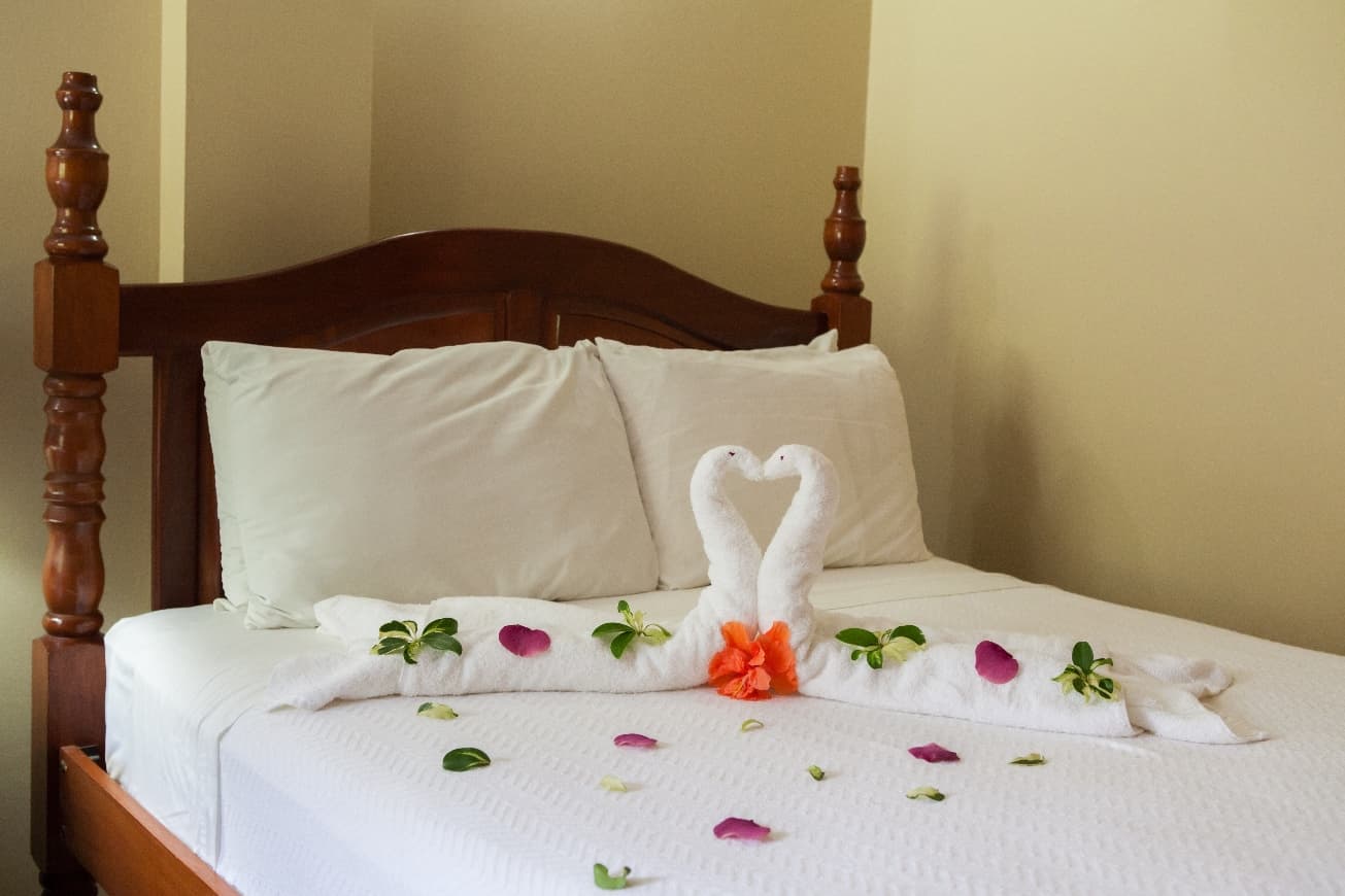 A hotel bed with white sheets and two towel swans arranged to form a heart, accented with rose petals and greenery.