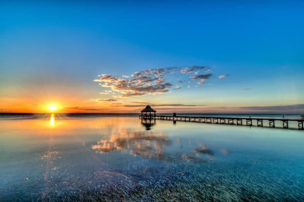 A vibrant sunset reflects on calm waters, with a pier leading to a gazebo.
