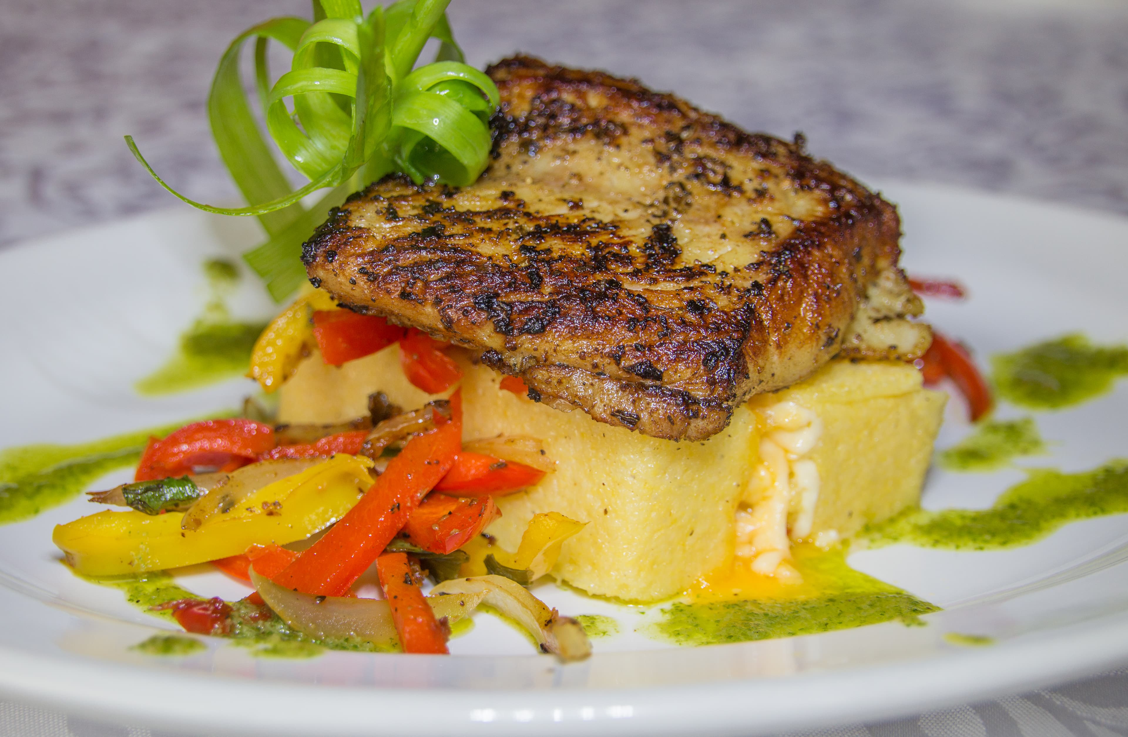 A beautifully plated dish featuring grilled fish atop creamy polenta, surrounded by colorful sautéed peppers and a drizzle of green sauce.