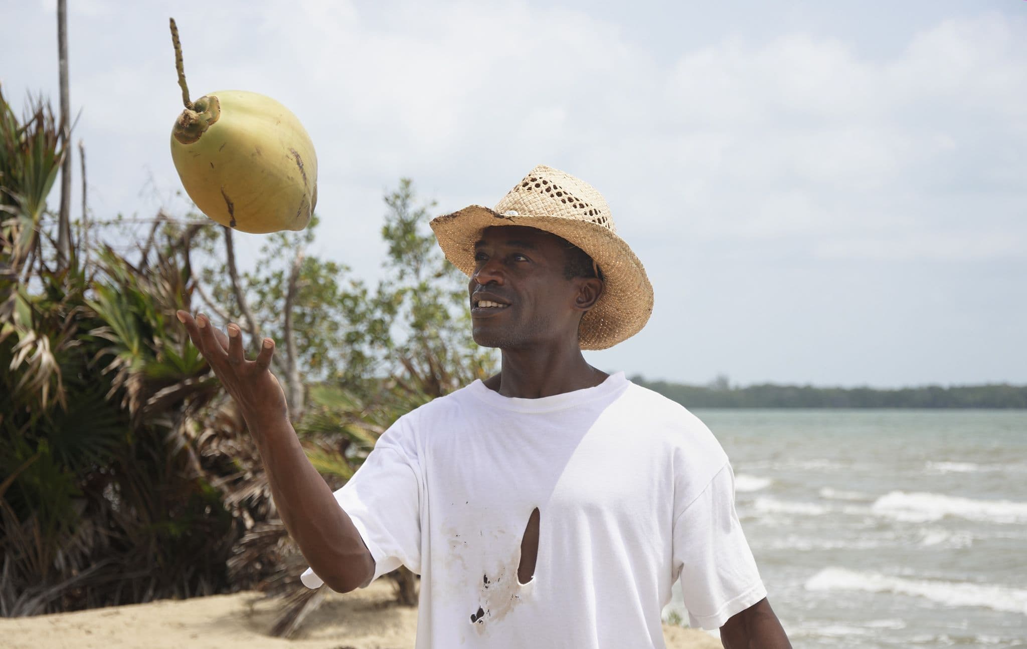 A man in a straw hat balances a coconut on his hand at the beach.