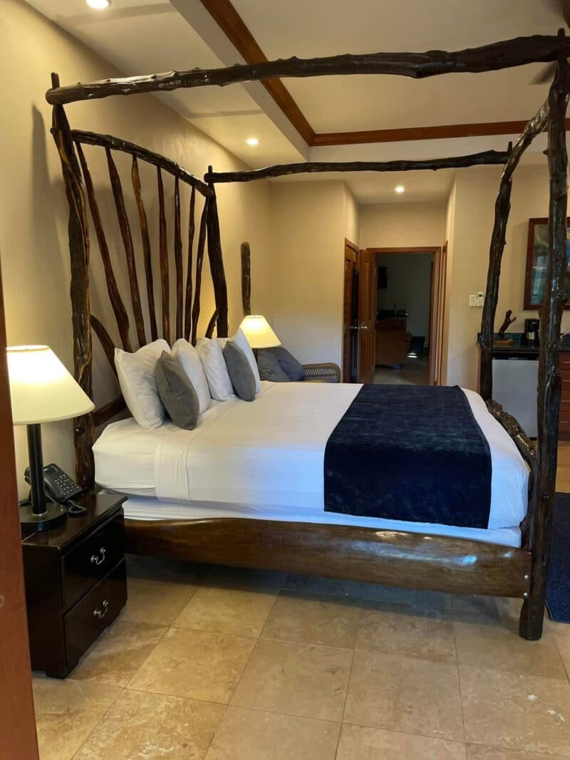 A cozy bedroom featuring a wooden canopy bed with decorative pillows, bedside lamps, and a doorway leading to another room.